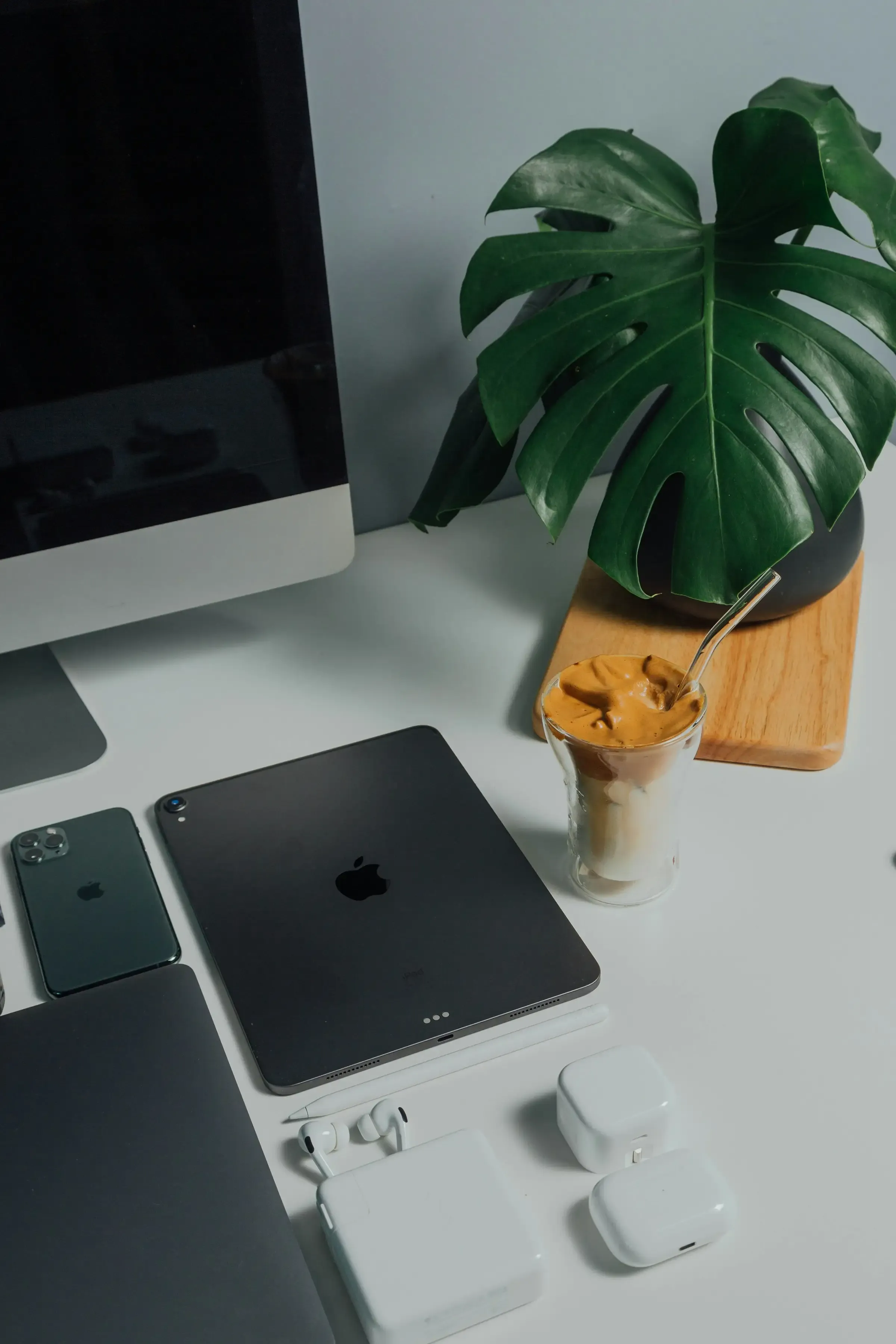A desk with a monitor, tablet, phone and earbuds laid out beside a monstera plant and an iced coffee.