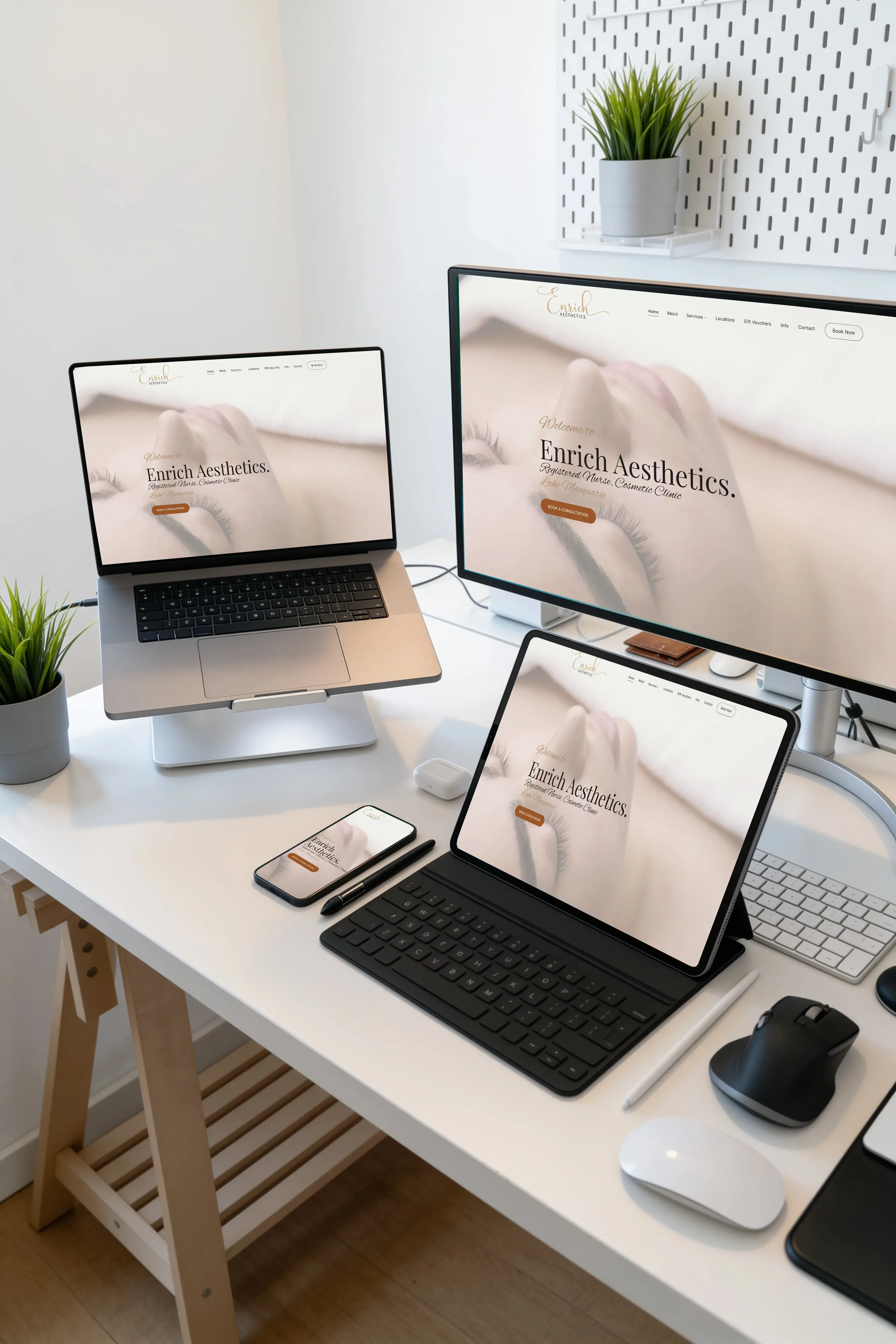 The Enrich Aesthetics home page running at once on a widescreen monitor, a laptop on a stand, a tablet in a keyboard case and a phone, laid out on a white desk.