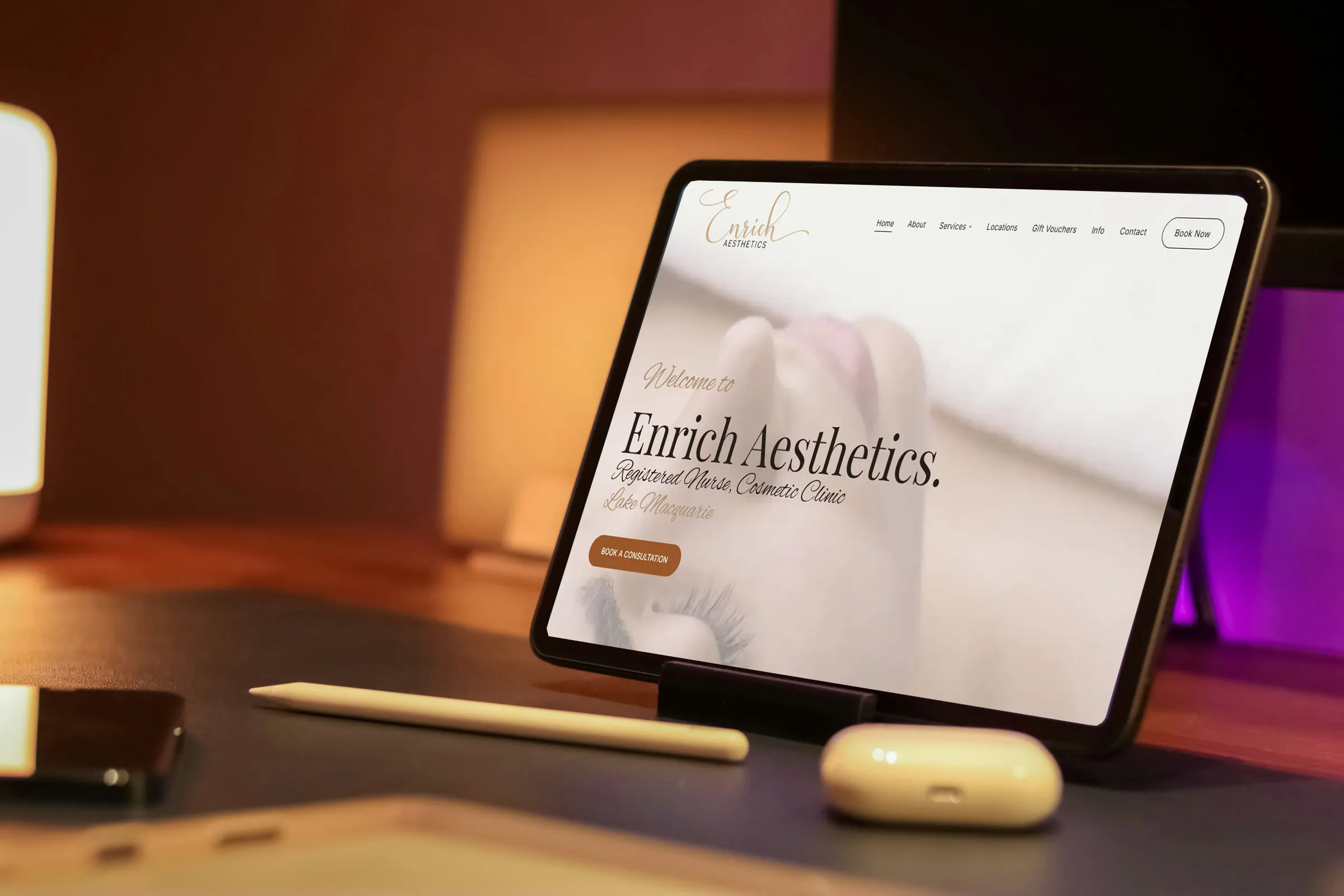 The Enrich Aesthetics home page on a tablet propped on a warmly lit desk, beside a stylus and earbuds.