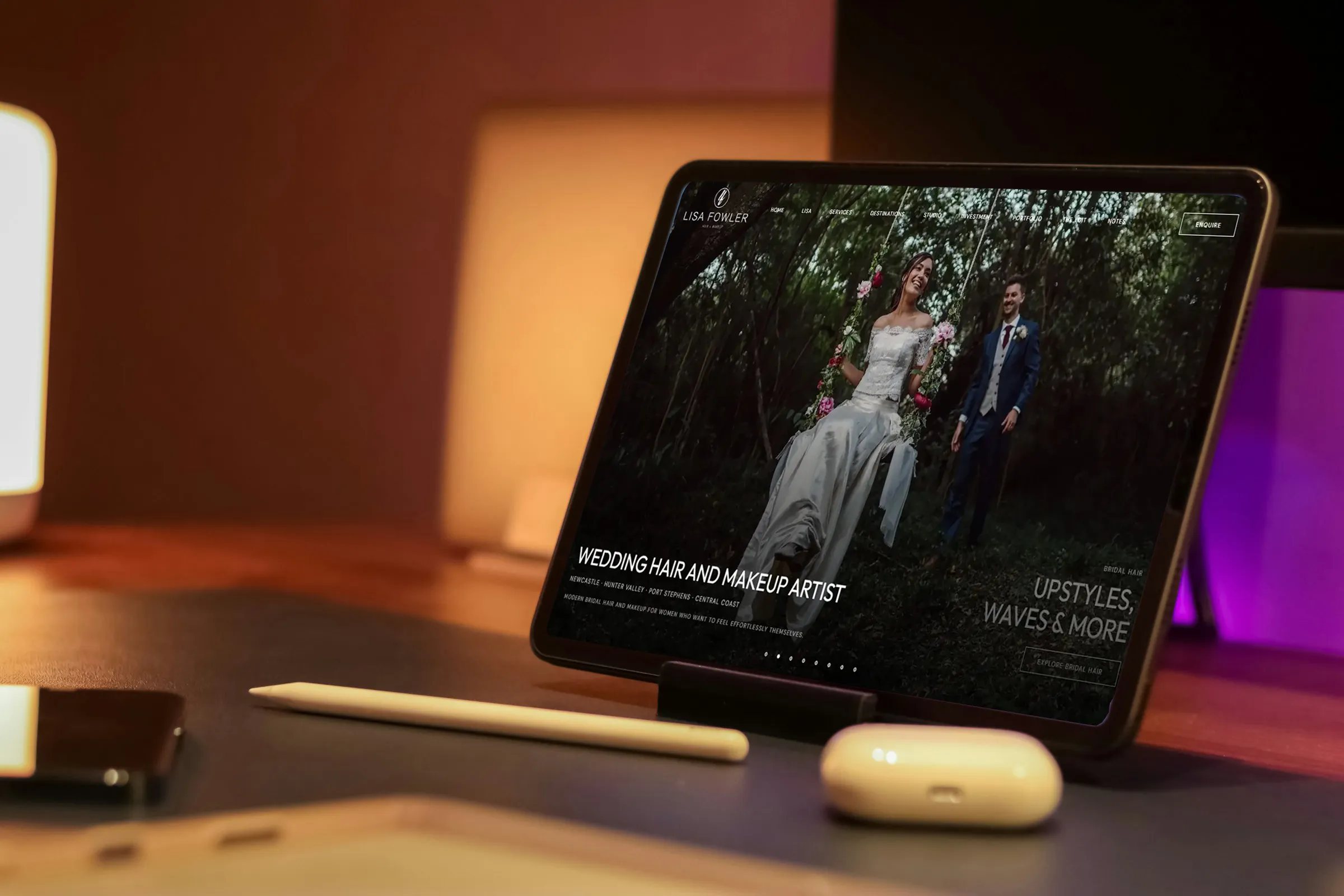 The Lisa Fowler Hair + Makeup home page on a tablet propped on a warmly lit desk, beside a stylus and earbuds.